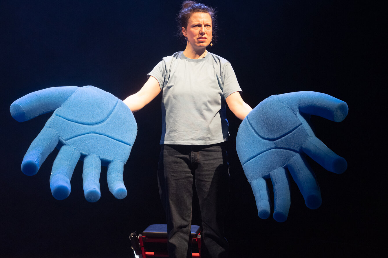 Eine Performerin auf einer Bühne hat ihre Hände einladend ausgestreckt. Sie trägt absurd große, blaue riesige Handschuhe. 