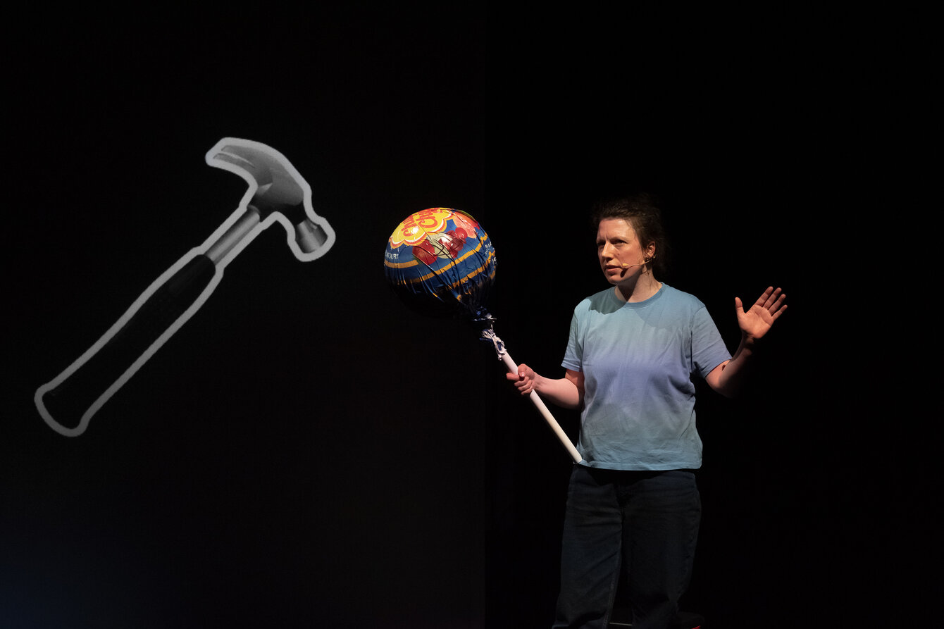 Eine Person in einem blauen T-Shirt ist steht auf einer dunklen Bühne. Sie hält einen übergroßen Lolli in der Hand, neben ihr ist eine große Projektion eines Hammers. 