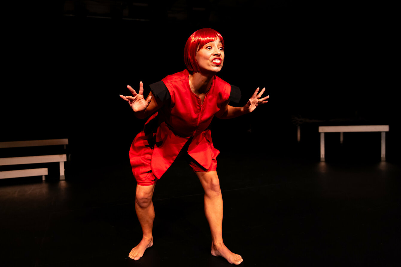 A scene from the performance subjoyride. The photo shows Boglárka Börcsök on a darkened stage. She has red hair and wears a bob with bangs and a red costume. She stands barefoot on the stage, her body slightly bent forward. Her hands are at chest height. With raised eyebrows, she looks forward to the right.