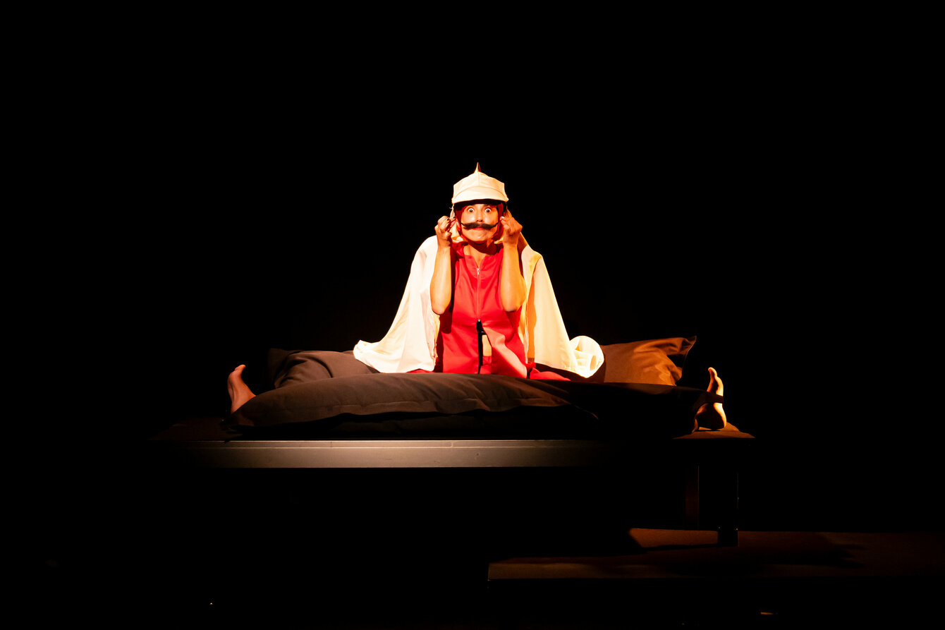 A scene from the performance subjoyride. The photo shows Boglárka Börcsök sitting barefoot with her legs spread apart on a darkened stage. She is wearing a red costume and a kind of white spiked helmet on her head. She has stuck on a moustache and is looking straight into the camera with her eyes wide open.  Her shoulders are covered by a white cape.