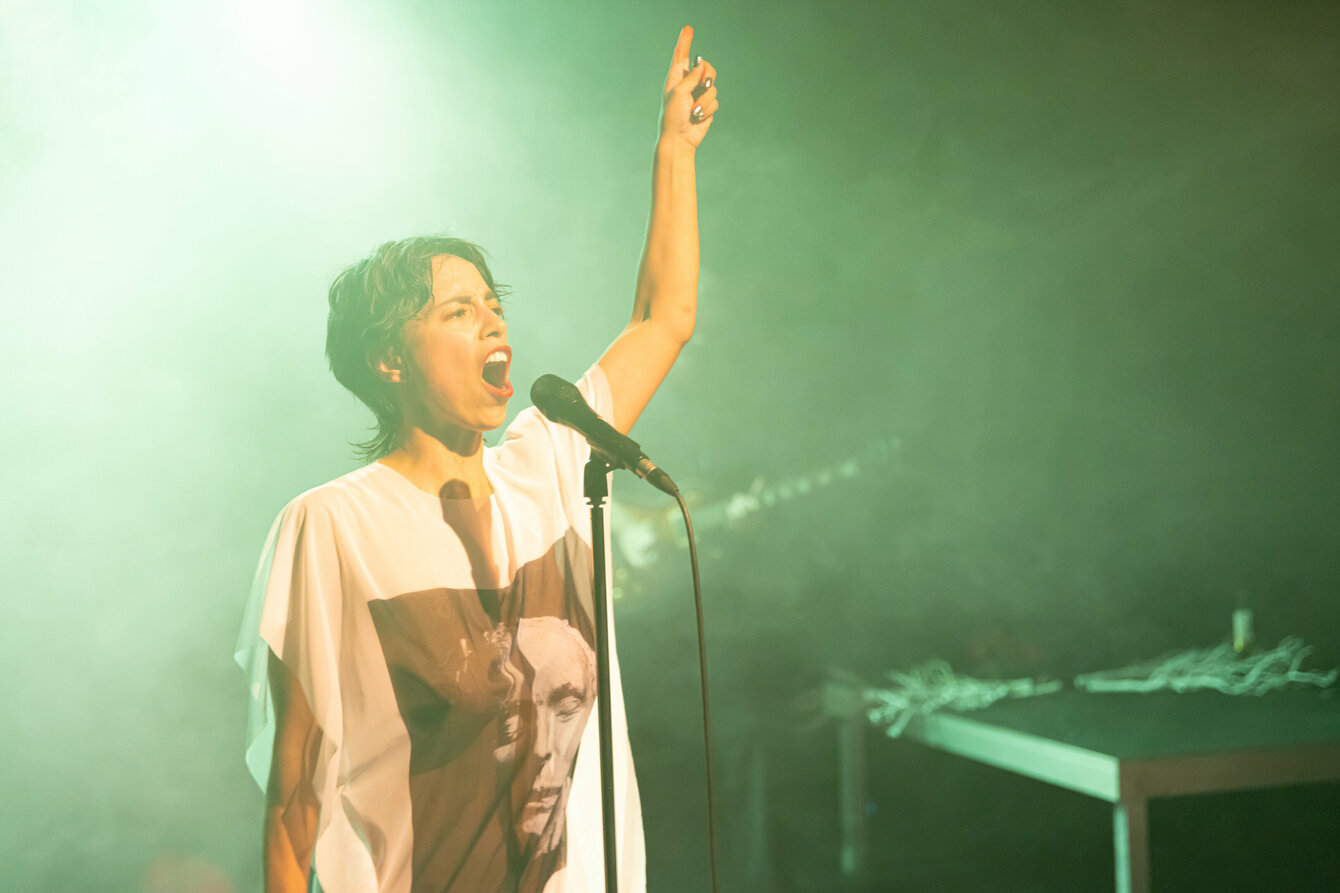 A scene from the performance subjoyride. The photo shows Boglárka Börcsök on a stage bathed in green light. She is standing behind a microphone, into which she appears to be singing. Her left arm is in the air, her index finger pointing upwards. Boglárka has short dark hair and is wearing a white shirt with a print showing a face.