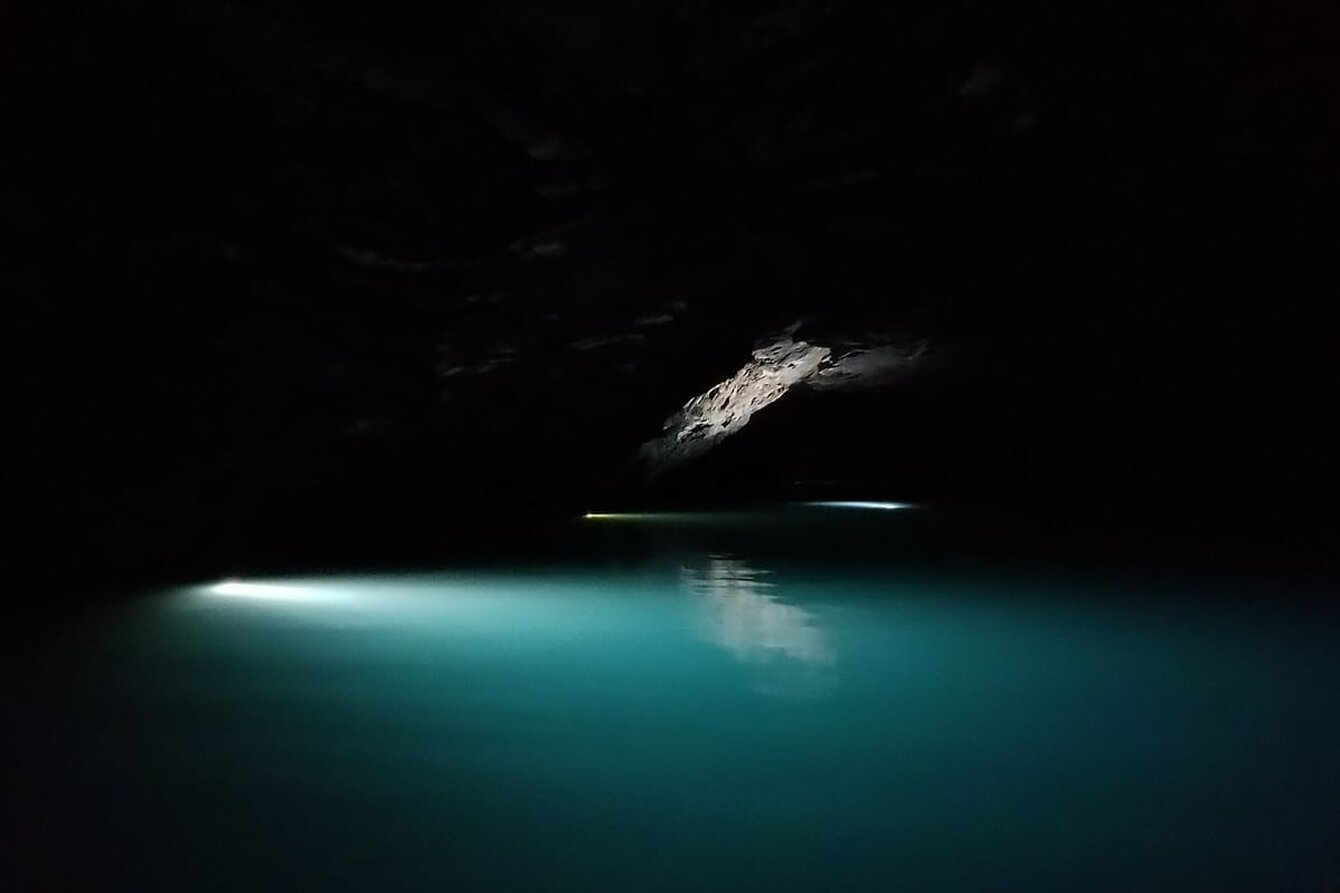 Ein unterirdischer See wird durch eine Höhlenöffnung leicht beschienen. Das Wasser sieht milchig aus und hat eine türkisene Färbung.