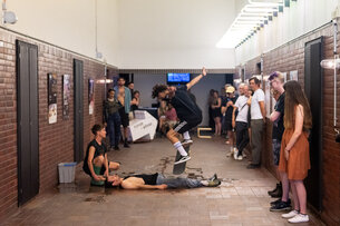 En Skater spring mit seinem Skateboard über eine liegende Person im Flur von PACT Zollverein