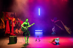 Eine Person steht auf einer Bühne. Sie trägt grüne Farbe am ganzen Körper. Sie spielt auf einer Gitarre und singt in ein Mikrofon. Im Hintergrund fährt eine Person auf Rollschuhen und zieht ein langes glitzerndes Gewand hinter sich her.