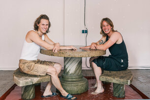 Gregor Leiprecht und Konstantin Pütz sitzen gut gelaunt und lachend im Kleinen Laden an ihrer interaktiven Installation. Diese besteht aus einem Wasserbecken, in welchem zwei steinerne Bänke und ein steinerner Tisch aufgestellt sind. Aus der Mitte des Tisches sprudelt aus einer Öffnung Wasser über und verteilt sich über die Fläche und das Sitzmobiliar, bevor es im Becken aufgefangen wird. 
