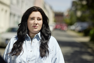 A portrait of Lena Schätte. Lena has long dark hair and is wearing a light blue shirt. She is standing on a street and looking at the camera.