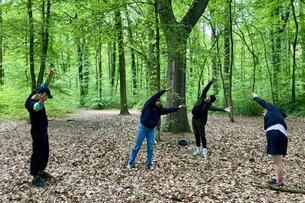 Four people interacting, performing and playing together in a forest. They stand at a distance from each other from left to right in the frame of the photograph, each striking a different pose.