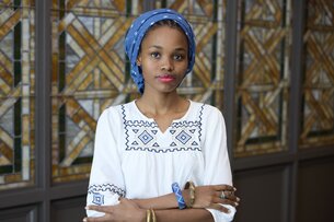 Portrait of Atang Arnold. Atang Arnold is wearing a blue headwear and a white top with with striking darker patterns. Atang Arnold looks directly into the camera with a bright and friendly expression, crossing the arms in front of the body.