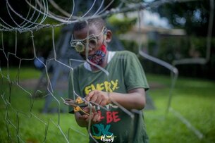 A black child is cutting a fence with a tool, wearing goggles and a face mask.