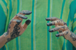 Two hands, painted with Henna are held towards the camera. The person in the background ist wearing a green and turquoise colored top.