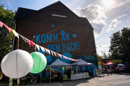 Food stalls, colourful flags and garlands are set up in front of a school building. The words ‘Come to the stream’ can be seen on the front of the building.