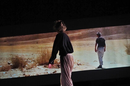 Eine Performerin steht auf einer Bühne im Dunkeln vor einer Videoprojektion. Auf dieser ist eine performende Person in selber Haltung zu sehen, welche sich jedoch in einer wüstenähnlichen Landschaft befindet. 