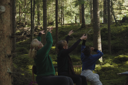 Drei Frauen sitzen auf Stühlen in einem Wald un bewegen ihre Hände