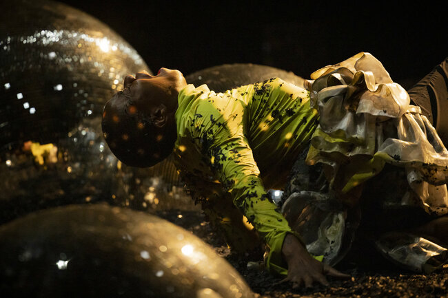 A scene from ‘Glitch Witch’: A performer dressed in green lies amid large glittering balls in a dark room. The person throws their head and body back and appears to be screaming. 