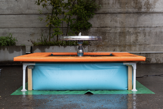 Eine Installation von Gregor Leiprecht. Es handelt sich um einen silbernen Brunnen im Form eines Tisches. Das Wasser fließt in ein Becken aus hellblauem Plastik. Unter dem Brunnen eine grüne Matte.