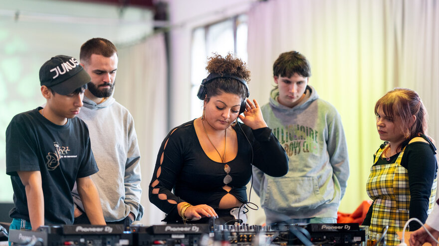 A group of young people stand in front of a DJ booth. One person is DJing, the others are watching closely as the person works.
