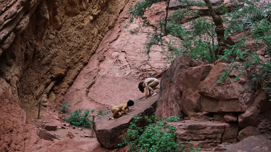 Zwei nackte Menschen, die mit Händen und Füßen auf einem Stein stehen. Sie schauen sich nicht an, sondern blicken nach unten. Hinter ihnen ein Felsenlandschaft.