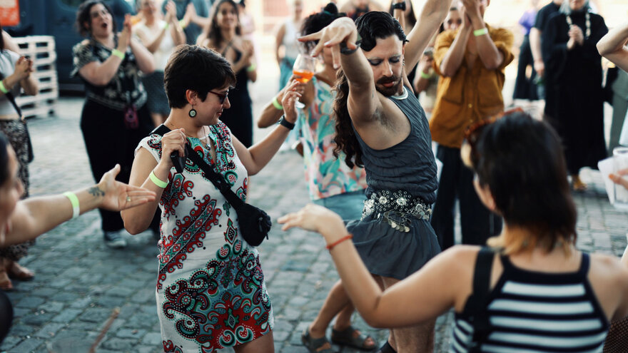 Eine Gruppe von Menschen tanzt gemeinsam im Freien auf Kopfsteinpflaster. Im Vordergrund hebt eine Person in einem grauen Kleid die Arme in einer expressiven Pose, während andere lachend und klatschend um sie herumstehen. Viele tragen bunte Kleidung und sommerliche Outfits, manche halten Getränke in der Hand. Die Stimmung wirkt ausgelassen, energiegeladen und gemeinschaftlich.