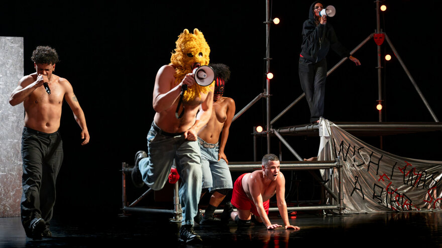 Eine Szene aus Delirious Night: Vier Performer*innen, oberkörperfrei, auf der Bühne, auf der ein Gerüst steht. Zwei von ihnen haben ein Mikro in der Hand. Im Bildvordergrund ein*e maskierte*r Performer*in mit Lautsprecher in der Hand.