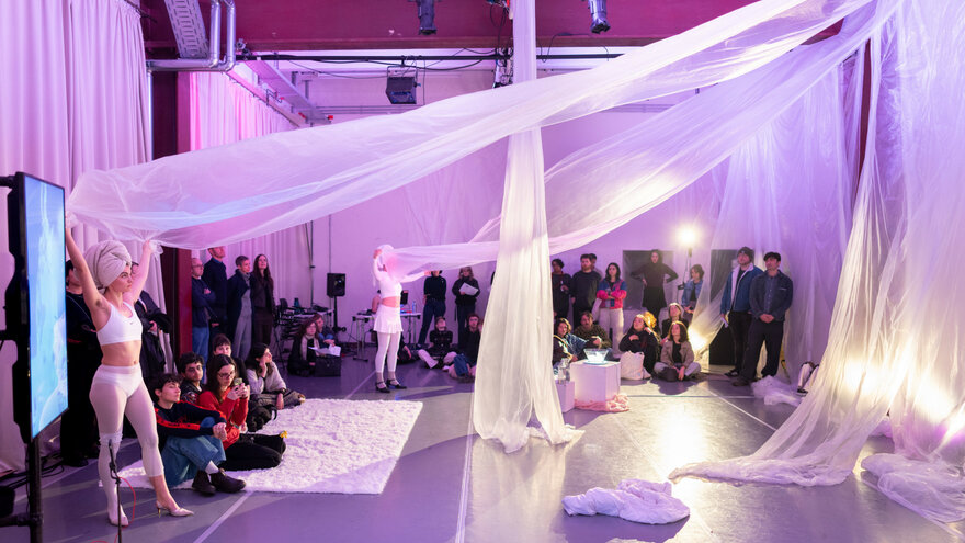 Eine Performance-Situation bei PACT Zollverein: Zwei Performer*innen, jeweils weiß gekleidet stehen auf der linken Seite des Raums, der pink-lila beleuchtet wird und halten weiße Stoffbahnen in der Hand, die oben an der gegenüberliegenden Wand angebracht sind. Das Publikum steht vor den Wänden.