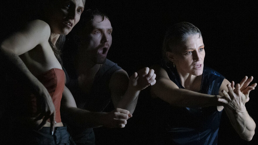 Eine Szene aus der Performance Mercury Rising: Drei Personen in dunkler Umgebung, die in einer dynamischen Pose stehen. Die Person links trägt ein ärmelloses, rotes Oberteil und eine lange, schwarze Hose. Die mittlere Person ist teilweise sichtbar und trägt ein dunkles Oberteil. Die Person rechts sitzt auf einem Hocker, trägt ein ärmelloses, dunkelblaues Oberteil und eine olivgrüne Hose, die Arme sind nach vorne ausgestreckt und die Hände ineinander verschränkt.