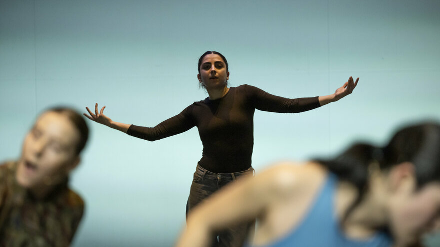 Das Foto zeigt drei Performer:innen auf einer Bühne, wobei zwei von ihnen nur angeschnitten und unscharf im Vordegrund zu erkennen sind. In der Bildmitte und im Fokus der Fotografie schaut eine Tänzer:in frontal in die Kamera streckt die Arme zur Seite aus. Der Hintergrund ist hell, die Performenden setzen sich deutlich von ihm ab. 