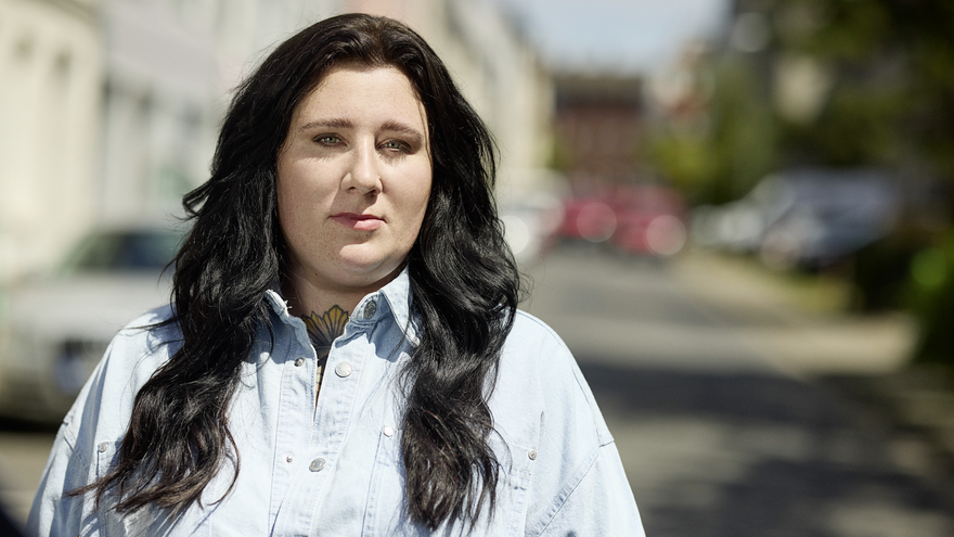 Ein Portrait von Lena Schätte. Lena hat lange dunkle Haare und trägt ein hellblaues Hemd. Sie steht auf einer Straße und blickt in die Kamera.