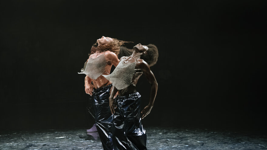 A scene from F*cking Future: Two performers can be seen dancing on stage. They both throw their heads back and push their chests forward. Both are wearing glittery silver sleeveless tops and black patent leather trousers.