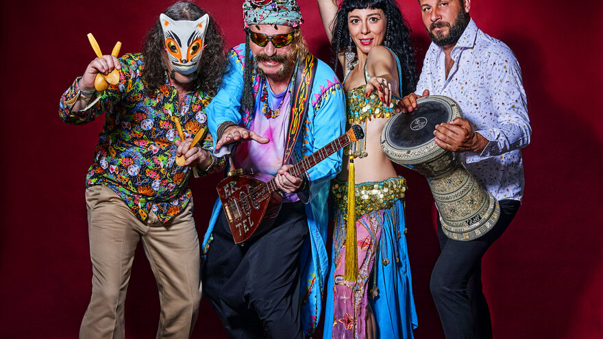Ein Gruppenfoto der Band Baba ZuLa: Vier bunt gekleidete Personen posieren stehend vor einer roten Wand. Links eine maskierte Person mit schulterlangen Haaren und dem Musikinstrument Löffel in der Hand, daneben Murat Ertel mit dem Instrument Electric Saz, daneben eine tanzende Esma Ertel und rechts neben ihr Perkussionist Ümit Adakale.