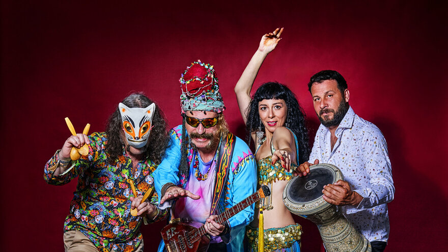 Ein Gruppenfoto der Band Baba ZuLa: Vier bunt gekleidete Personen posieren stehend vor einer roten Wand. Links eine maskierte Person mit schulterlangen Haaren und dem Musikinstrument Löffel in der Hand, daneben Murat Ertel mit dem Instrument Electric Saz, daneben eine tanzende Esma Ertel und rechts neben ihr Perkussionist Ümit Adakale.