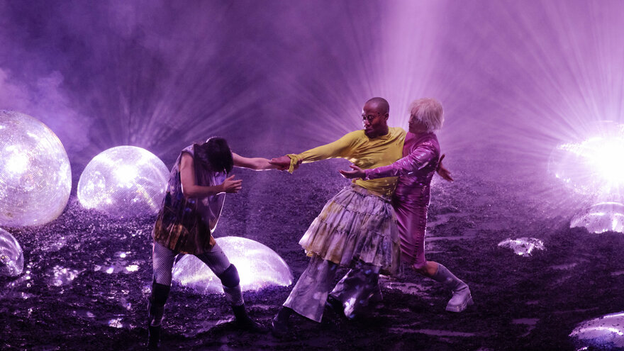 Eine Szene aus der Performance GLITCH WITCH. In der Bildmitte drei Performer*innen auf einer Bühne, die in lila Licht getaucht ist. Auf dem Bühnenboden sind riesige, leuchtende Diskokugeln zu sehen