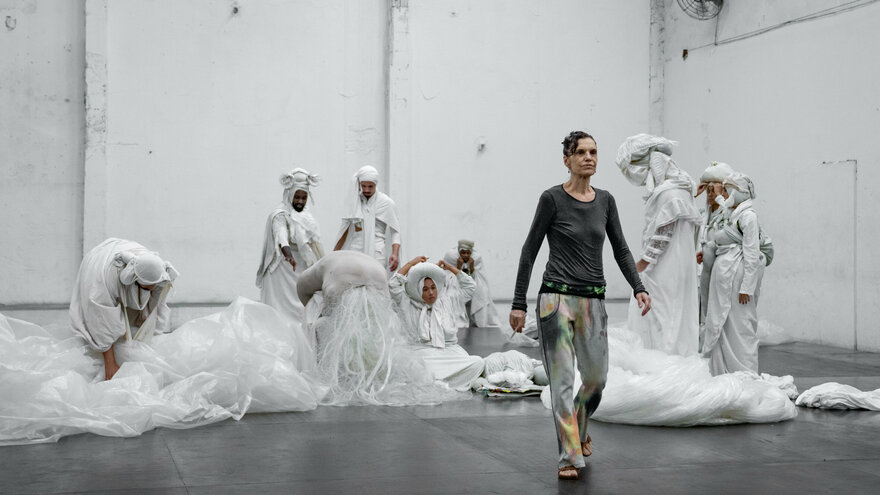 A hall, in it the choreographer Lia Rodrigues and the performers of her piece “BORDA”. Lia Rodrigues is wearing a dark top and gray trousers with a colour gradient. She walks upright to the front. Behind her are the performers, their bodies wrapped in white fabrics. They are preoccupied with themselves.  