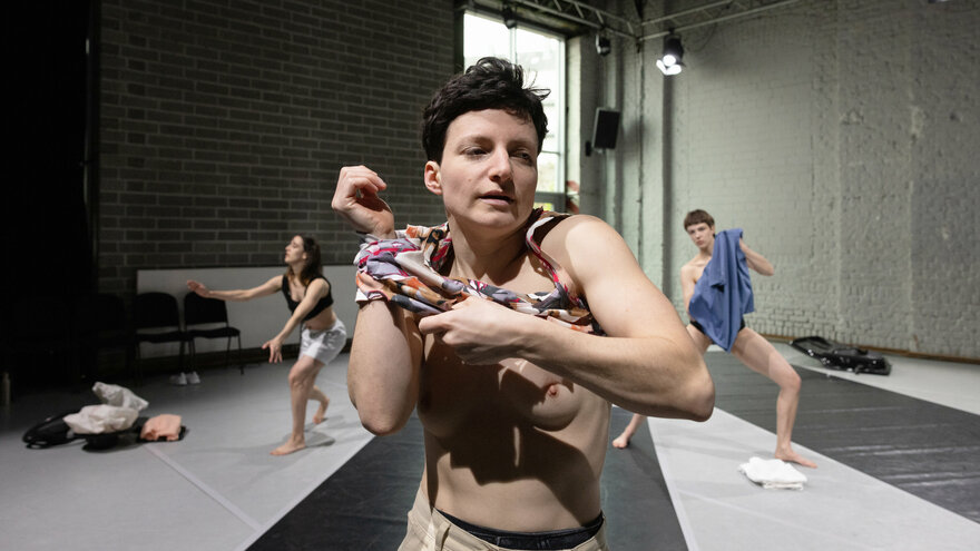Das Bild zeigt eine weiblich gelesene Performerin, die sich in einem Tanzstudio ihr Hemd auszieht. Im Hintergrund sind zwei weitere Tänzer:innen zu sehen. Der Raum hat Backsteinwände und große Fenster. Der Tanzboden hat breite, dunkle Streifen.