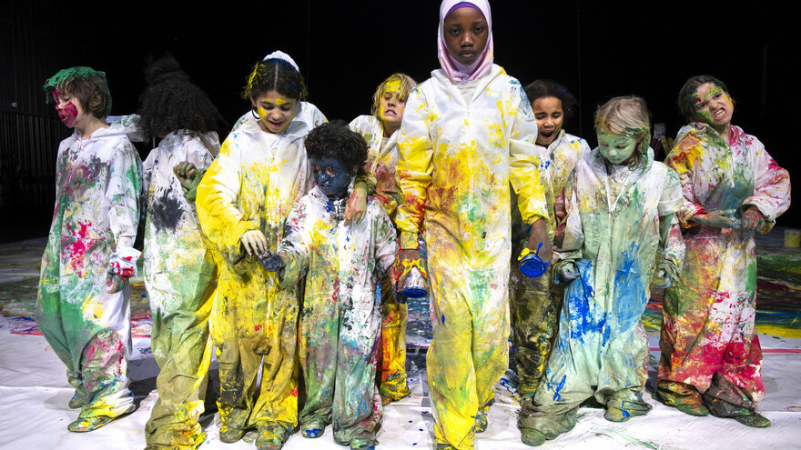 A group of children run towards the camera. They are all wearing painter's overalls that are smeared with lots of colors. Some of the children have paint on their faces. 